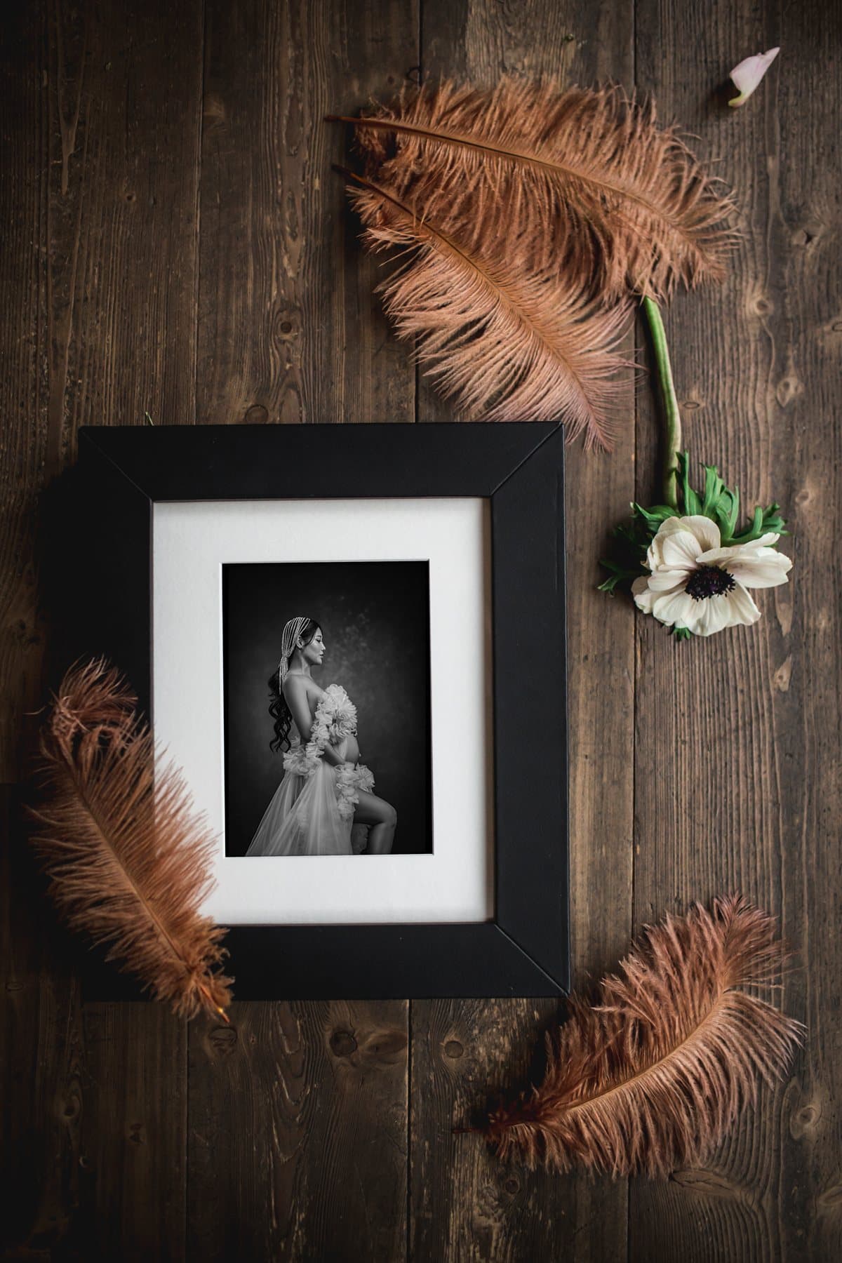 A framed fine art boudoir print styled for a reveal, with ostrich feathers and a white anemone on dark wood